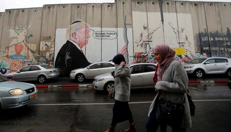 فلسطينيات يمشين بجوار جدار العزل الإسرائيلي عليه صورة ترامب- (رويترز)
