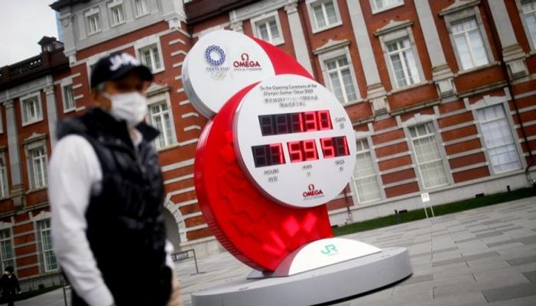 رجل يرتدي كمامة أمام ساعة عملاقة للألعاب الأولمبية بطوكيو 2020