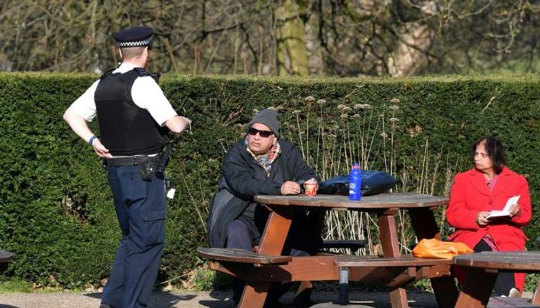 شرطي بريطاني يتحدث إلى زوجين جالسين على مقعد في متنزه 