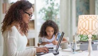 سيدة تعمل على الحاسوب في منزلها وبجوارها طفلة تدرس